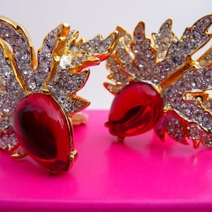 May include: A pair of gold-toned earrings with a red teardrop-shaped center stone. The earrings feature a detailed design with clear crystals and a gold-colored metal frame, set against a bright pink background. The earrings have a floral or leaf-like design.