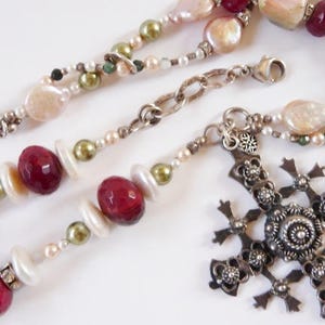 May include: A necklace featuring a silver-toned metal cross pendant with intricate filigree details. The chain consists of alternating red faceted beads, iridescent white pearls, and small olive green beads, connected by silver-toned metal links.