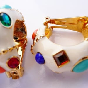 May include: A pair of gold-tone hoop clip-on earrings featuring a smooth white enamel finish. Each earring is decorated with an assortment of colorful, multi-shaped cabochon and faceted stones in shades of blue, green, red, and orange.