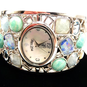 May include: Silver-tone cuff watch with an oval face and a mother-of-pearl dial. The watch is adorned with various colorful gemstones in shades of blue, green, and white. The watch face displays the brand name "Victoria Pink" and the numbers 3, 6, and 12.