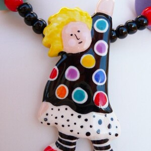 May include: A ceramic pendant shaped like a figure with yellow hair, wearing a black top with colorful polka dots and a white skirt with black dots. The figure has striped legs and red shoes, suspended on a necklace of black, red, and teal beads.