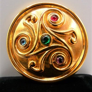 May include: A circular gold-toned brooch featuring a raised, swirling triskelion design. The metal surface is polished and smooth, accented with four small, round, cabochon-style gemstones in red, green, blue, and purple, set within the spirals.