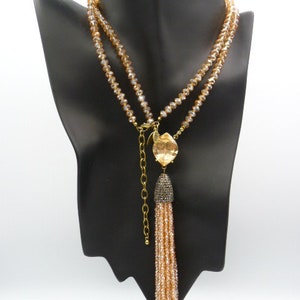 May include: A multi-strand necklace featuring light champagne-colored faceted crystal beads. The design includes a central gold-toned metal setting holding a large, clear, marquise-shaped crystal pendant attached to a long, beaded tassel with a dark cap.