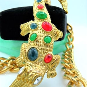 May include: A gold-toned alligator brooch with colorful gemstone accents. The brooch features red, green, and blue stones, and clear crystals on the snout. A gold chain is visible in the background. This vintage-style jewelry piece is a statement accessory.