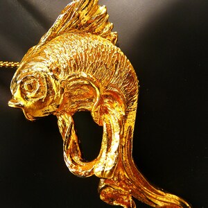 May include: A gold-colored fish pendant on a gold chain. The pendant is intricately detailed, with textured scales and fins. The fish is stylized with a flowing tail and a loop in its body. The background is black.