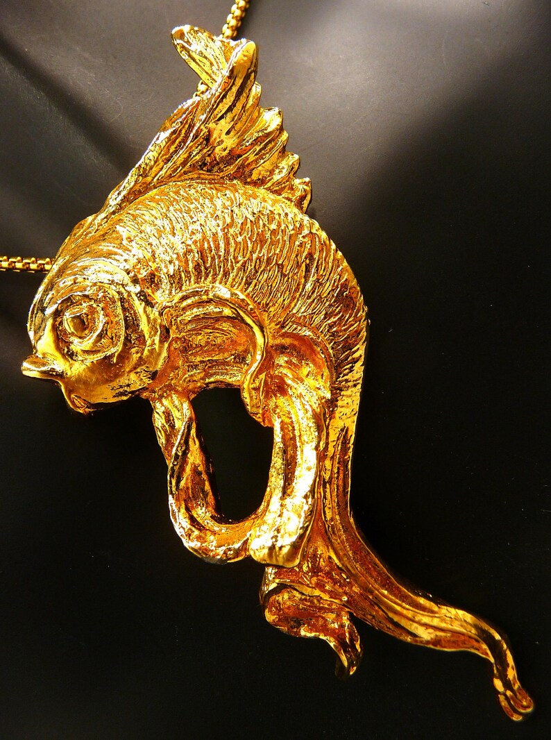 May include: A gold-colored fish pendant on a gold chain. The pendant is intricately detailed, with textured scales and fins. The fish is stylized with a flowing tail and a loop in its body. The background is black.