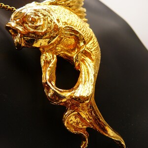May include: A gold-colored fish pendant with intricate details, including scales and fins. The pendant is suspended from a delicate chain. The fish is sculpted with an open mouth and flowing tail, creating a dynamic and elegant design.