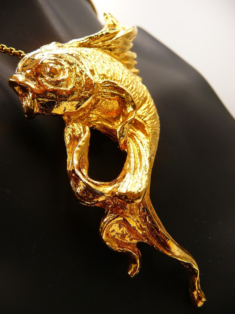 May include: A gold-colored fish pendant with intricate details, including scales and fins. The pendant is suspended from a delicate chain. The fish is sculpted with an open mouth and flowing tail, creating a dynamic and elegant design.