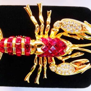 May include: A gold-toned lobster brooch with red and clear crystal accents. The lobster's body is composed of red rectangular crystals, while the claws and tail feature clear crystals. The brooch is set against a black background.