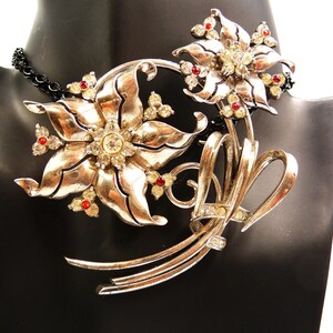 May include: A silver-toned floral necklace with red and clear crystal accents. The necklace features two large flower pendants and a delicate chain. The flowers have detailed petals and are adorned with sparkling crystals, creating a vintage-inspired look.