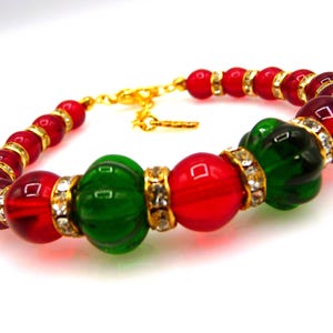 May include: A beaded bracelet featuring alternating round red and green glass beads, accented with gold-toned spacers embedded with small clear crystals. The bracelet includes a gold-toned metal chain clasp and a small gold-toned metal tag.