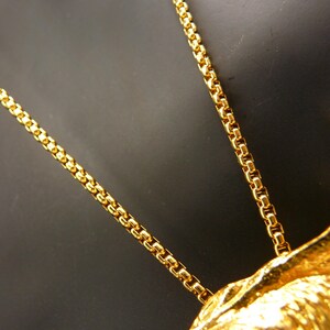 May include: Close-up of a gold-colored necklace with a detailed chain design. The necklace is set against a dark background, highlighting the intricate details of the chain and pendant. The chain is made of small, rectangular links.