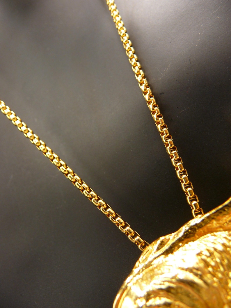 May include: Close-up of a gold-colored necklace with a detailed chain design. The necklace is set against a dark background, highlighting the intricate details of the chain and pendant. The chain is made of small, rectangular links.