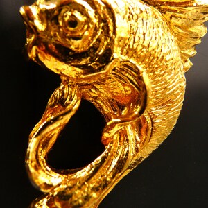 May include: A close-up of a gold-colored fish pendant. The pendant is intricately detailed, with textured scales and fins. The fish is suspended from a delicate gold chain, creating a striking piece of jewelry.