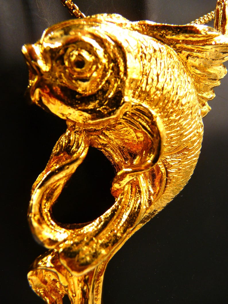 May include: A close-up of a gold-colored fish pendant. The pendant is intricately detailed, with textured scales and fins. The fish is suspended from a delicate gold chain, creating a striking piece of jewelry.