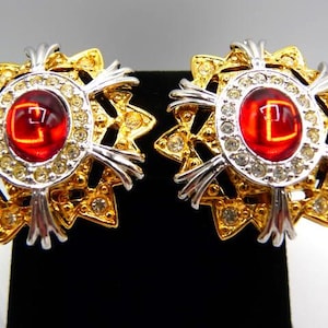 May include: A pair of ornate earrings with a gold and silver design. Each earring features a central red stone surrounded by clear crystals and a gold starburst pattern. The earrings are displayed on a black surface.