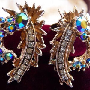 May include: A pair of gold-tone metal earrings featuring a curved, leaf-like design adorned with iridescent blue and green round crystals and a row of clear, sparkling rhinestones along the inner edge, creating a textured, multi-faceted appearance.