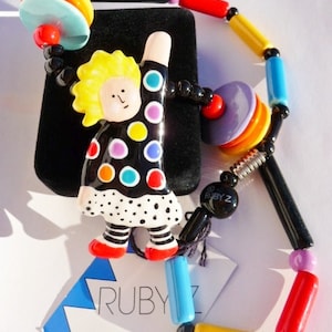 May include: A colorful necklace featuring a whimsical figure with a yellow hairstyle and a black dress with polka dots. The necklace has a variety of beads in red, yellow, blue, and black. The tag reads "RUBY Z".