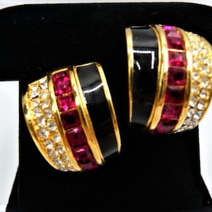 May include: A pair of gold-tone hoop earrings featuring a wide band design. Each earring is decorated with a central row of square-cut magenta crystals, a smooth black enamel stripe, and an outer edge of small, clear round rhinestones.