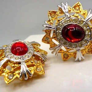 May include: A pair of circular earrings featuring a central oval red cabochon stone surrounded by a ring of small clear crystals. The base is a gold-tone metal starburst design with silver-tone accents and additional small clear crystals.