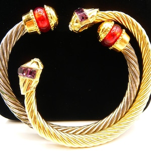 May include: A bypass cuff bracelet featuring two twisted metal bands, one in silver tone and one in gold tone. The ends are capped with gold tone metal accents, two featuring round red glass beads and two featuring rectangular purple glass stones.