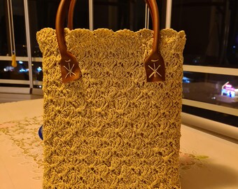 Puede incluir: Bolso de mano tejido a ganchillo en tono amarillo claro, con asas marrones y detalles decorativos. El bolso tiene un adorno floral en la parte superior y un patrón texturizado. El bolso mide unos 25 cm de alto.