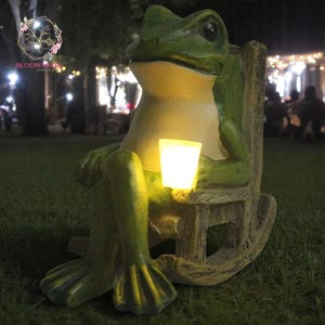 Può includere: Una statua di rana verde seduta su una sedia a dondolo, che tiene in mano una lanterna luminosa. La rana ha un corpo verde chiaro e una testa verde più scura. La sedia è di colore marrone chiaro. La lanterna emette una luce gialla calda.