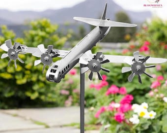 May include: A silver metal aeroplane wind spinner with four propellers. The aeroplane is mounted on a tall metal pole. The wind spinner is in a garden setting with flowers in the background.