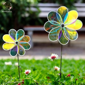 Puede incluir: Dos molinetes de viento con forma de flor iridiscente con un efecto arcoíris. El molinete más grande está en el fondo y el más pequeño en primer plano. Ambos molinetes están en estacas de metal y se encuentran en un área cubierta de hierba con flores blancas.