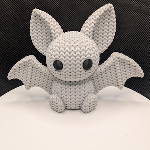 Può includere: Una statuetta di pipistrello grigio chiaro con una superficie testurizzata simile alla maglia. Il pipistrello ha grandi orecchie a punta, ali spiegate e due occhi neri lisci e rotondi. La figura è raffigurata in posizione seduta.