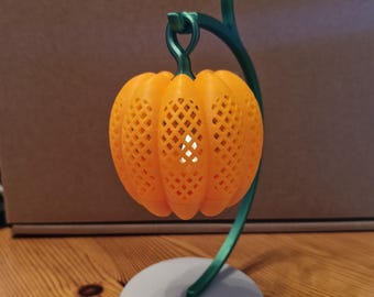 May include: An orange, 3D-printed pumpkin-shaped lantern with a lattice design, suspended from a green, curved stem. The lantern sits on a gray, circular base. The design is suitable for Halloween or autumn decor.