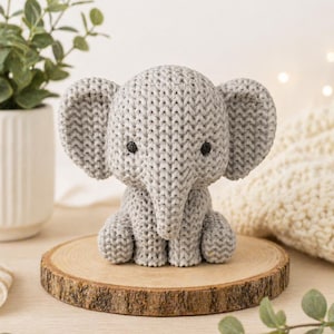 Puede incluir: Un juguete de elefante gris tejido con grandes orejas y pequeños ojos negros. El elefante está sentado sobre una rodaja de madera redonda. El fondo incluye una maceta blanca con vegetación y una manta suave blanquecina.