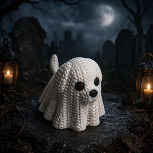 Op de afbeelding: Een handgemaakte witte gebreide spookhond met zwarte ogen en neus. De spookhond staat tegen een griezelige kerkhofscène met een volle maan en verlichte lantaarns, perfect voor Halloween-decoraties.