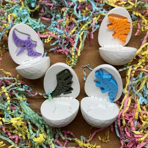 May include: Four plastic dinosaur eggs with different colored dinosaurs inside. The eggs are white with a speckled pattern and open to reveal the dinosaurs.