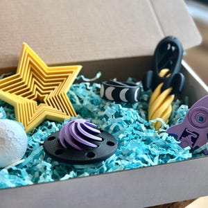 May include: A gift box filled with blue tissue paper and a variety of 3D printed space-themed toys, including a purple rocket, a yellow star, a black and white ring, a purple and black swirl, and a white moon.