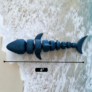 May include: A blue plastic toy shark, segmented into multiple pieces, measuring 6 inches in length. The shark is lying on a white sandy beach with foamy waves.