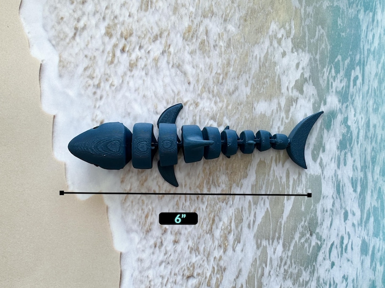 May include: A blue plastic toy shark, segmented into multiple pieces, measuring 6 inches in length. The shark is lying on a white sandy beach with foamy waves.