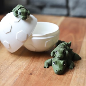 May include: A white, egg-shaped container with a lid, decorated with hexagonal patterns. Two green alligator figurines are placed on the lid and beside the container. The container is open, revealing its interior.