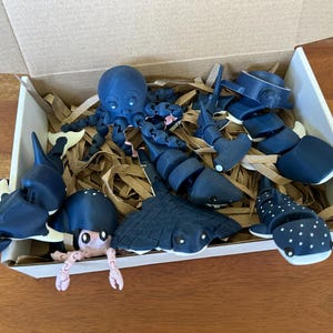 May include: A box filled with blue and white 3D printed sea creatures, including an octopus, a stingray, a shark, a whale shark, and a seahorse. The creatures are arranged in a bed of brown packing paper.
