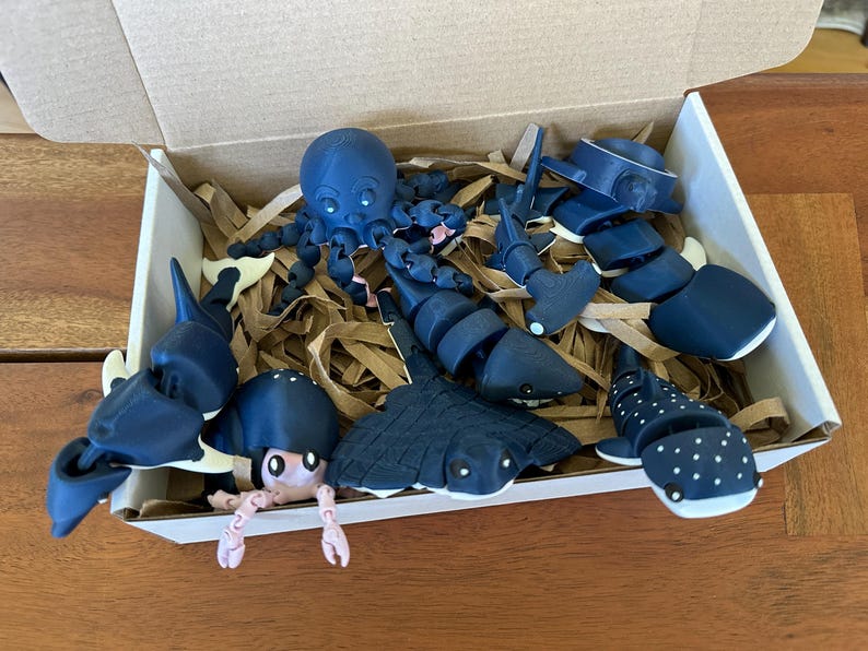 May include: A box filled with blue and white 3D printed sea creatures, including an octopus, a stingray, a shark, a whale shark, and a seahorse. The creatures are arranged in a bed of brown packing paper.