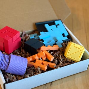 May include: A variety of colorful 3D puzzles and shapes are arranged inside a white box with brown packing material. The puzzles include a red cube, a black block, orange geometric shapes, a blue puzzle, a yellow cube, and a purple cylinder.
