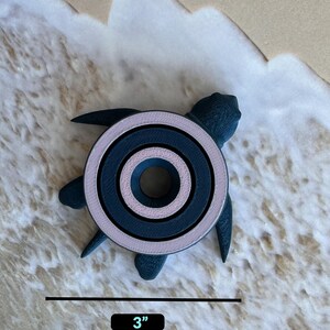 May include: A dark blue and pink turtle-shaped ring holder with three concentric circles. The holder is 3 inches in diameter and is sitting on a sandy surface.