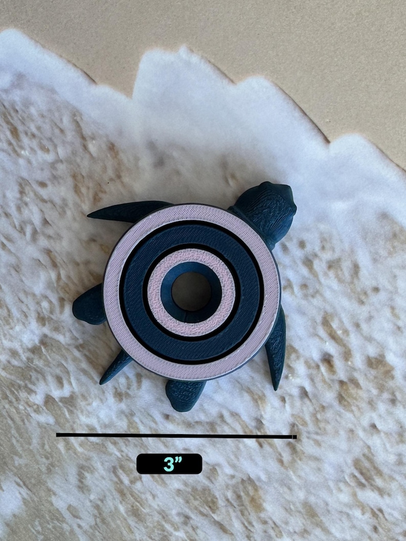 May include: A dark blue and pink turtle-shaped ring holder with three concentric circles. The holder is 3 inches in diameter and is sitting on a sandy surface.