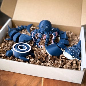 May include: A set of blue and white ocean animal figurines in a cardboard box with brown packing paper. The set includes a shark, an octopus, a turtle, a whale shark, and a manta ray.