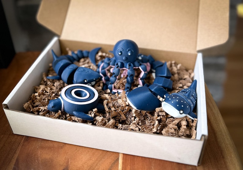 May include: A set of blue and white ocean animal figurines in a cardboard box with brown packing paper. The set includes a shark, an octopus, a turtle, a whale shark, and a manta ray.
