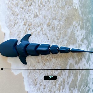 May include: A dark blue articulated shark toy, measuring 7 inches long. The toy is made of plastic and has a segmented body.