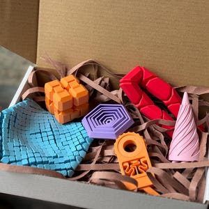May include: A cardboard box filled with colorful 3D-printed geometric shapes. The shapes include an orange cube, a red articulated form, a purple hexagon, a blue square, an orange tool, and a pink cone. The box is filled with brown paper shreds.