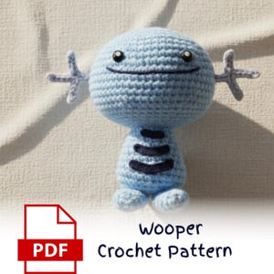 May include: A light blue crochet Wooper doll with dark blue accents and a smiling face. The doll has small arms and feet. The text "Wooper Crochet Pattern" is below the doll, along with a PDF icon.