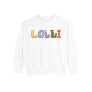 Peut inclure: Un sweat-shirt blanc à manches longues avec le mot LOLLI sur la poitrine. Chaque lettre est représentée dans une couleur et un motif différents, notamment du vichy jaune, des pois bleus, des fleurs violettes, des points verts et un imprimé floral rouge.