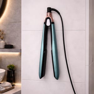 Puede incluir: Una plancha de pelo turquesa y negra con detalles en oro rosa cuelga de una pared de azulejos blancos. La plancha tiene un cable de alimentación negro. El entorno es un baño moderno.